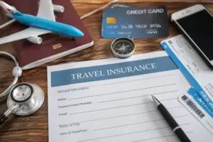 ภาพแนวสมจริงเกี่ยวกับประกันการเดินทาง มีแบบฟอร์ม Travel Insurance พร้อมพาสปอร์ต เครื่องบินจำลอง บัตรเครดิต ตั๋วเครื่องบิน สมาร์ตโฟน และหูฟังแพทย์วางบนโต๊ะไม้ สื่อถึงการวางแผนท่องเที่ยวและความคุ้มครองระหว่างเดินทาง